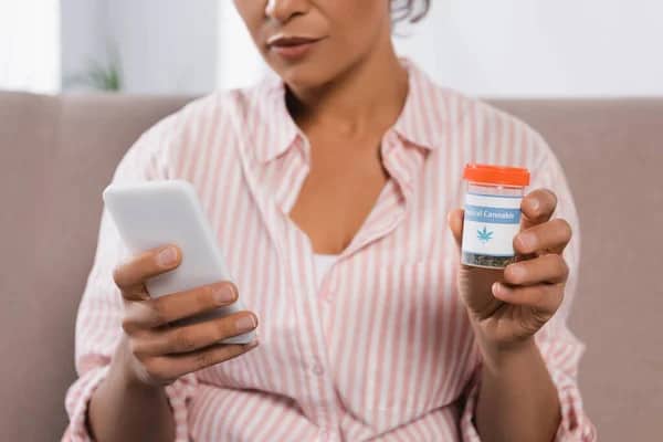 woman holding phone in one hand and bottle of medical cannabis buds in the other